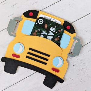 May include: A yellow school bus-shaped gift card holder with black wheels and accents. The bus has blue windows, a red stop light, and a gift card that says "THANK YOU" and "Holidays".