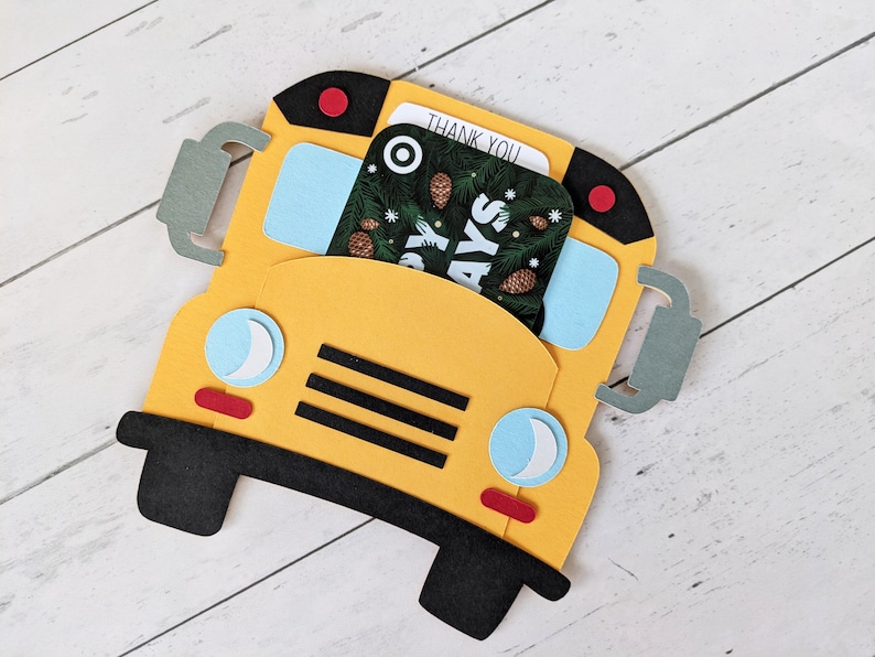 May include: A yellow school bus-shaped gift card holder with black wheels and accents. The bus has blue windows, a red stop light, and a gift card that says "THANK YOU" and "Holidays".