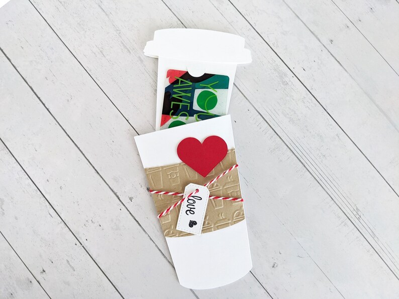 Puede incluir: Un vaso de caf&eacute; de papel blanco con una tarjeta de regalo dentro. El vaso est&aacute; decorado con un coraz&oacute;n rojo y una etiqueta que dice "love".
