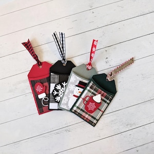 Puede incluir: Cuatro etiquetas de regalo navide&ntilde;as con cintas decorativas. Cada etiqueta presenta un dise&ntilde;o diferente: un guante, un copo de nieve, un mu&ntilde;eco de nieve y una taza de chocolate caliente. Las etiquetas son rojas, negras, grises y verdes con detalles a cuadros.
