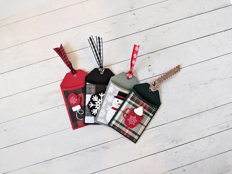 Puede incluir: Cuatro etiquetas de regalo navide&ntilde;as con cintas decorativas. Cada etiqueta presenta un dise&ntilde;o diferente: un guante, un copo de nieve, un mu&ntilde;eco de nieve y una taza de chocolate caliente. Las etiquetas son rojas, negras, grises y verdes con detalles a cuadros.