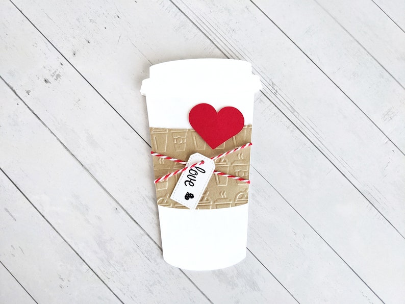 Puede incluir: Un vaso de caf&eacute; de papel blanco con una manga de papel marr&oacute;n decorada con vasos de caf&eacute; y vapor. El vaso est&aacute; atado con una cuerda roja y blanca y tiene un coraz&oacute;n rojo y una etiqueta que dice "love" adjunta.