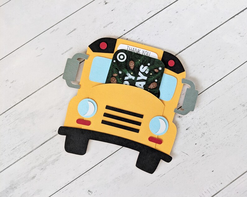 May include: A yellow school bus-shaped card holder with black wheels and accents. The bus has blue windows, red lights, and grey side mirrors. A card with a pine cone design and the words "THANK YOU" and "HAPPY DAYS" is inserted into the holder.