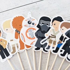 Puede incluir: Un conjunto de 10 adornos para cupcakes de personajes de Star Wars. Los adornos presentan personajes como Darth Vader, Chewbacca, Rey y Finn. Los adornos est&aacute;n impresos en cartulina blanca y unidos a brochetas de madera.
