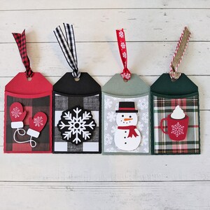 Puede incluir: Cuatro etiquetas de regalo navide&ntilde;as con elementos decorativos. Las etiquetas presentan fondos rojos, negros, verdes y grises con dise&ntilde;os festivos. Cada etiqueta tiene una cinta para colgar. Los dise&ntilde;os incluyen mitones, un copo de nieve, un mu&ntilde;eco de nieve y una taza.
