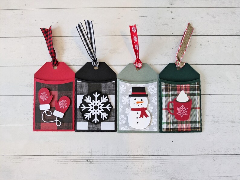 Puede incluir: Cuatro etiquetas de regalo navide&ntilde;as con elementos decorativos. Las etiquetas presentan fondos rojos, negros, verdes y grises con dise&ntilde;os festivos. Cada etiqueta tiene una cinta para colgar. Los dise&ntilde;os incluyen mitones, un copo de nieve, un mu&ntilde;eco de nieve y una taza.