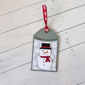 Puede incluir: Una etiqueta de regalo hecha a mano con un dise&ntilde;o de mu&ntilde;eco de nieve. La etiqueta tiene un fondo gris con un patr&oacute;n de copos de nieve blancos. El mu&ntilde;eco de nieve tiene un sombrero negro, una nariz naranja y una bufanda roja. Se adjunta una cinta roja con copos de nieve blancos.