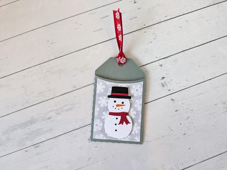 Puede incluir: Una etiqueta de regalo hecha a mano con un dise&ntilde;o de mu&ntilde;eco de nieve. La etiqueta tiene un fondo gris con un patr&oacute;n de copos de nieve blancos. El mu&ntilde;eco de nieve tiene un sombrero negro, una nariz naranja y una bufanda roja. Se adjunta una cinta roja con copos de nieve blancos.