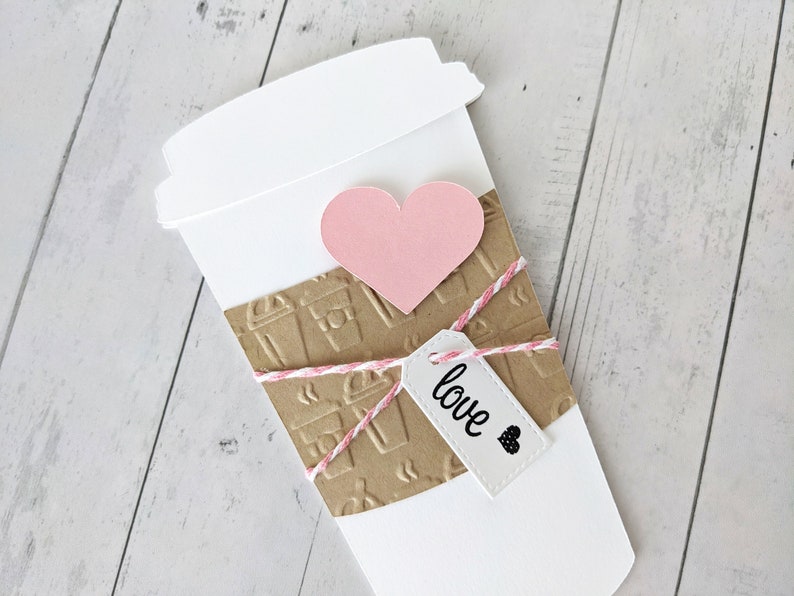 Puede incluir: Un vaso de caf&eacute; de papel blanco con una manga de papel marr&oacute;n decorada con iconos de vasos de caf&eacute;. Un coraz&oacute;n rosa est&aacute; unido al vaso y una etiqueta blanca con la palabra "love" est&aacute; atada al vaso con una cuerda.