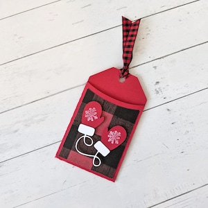 Puede incluir: Una etiqueta de regalo roja con un patr&oacute;n de cuadros negros y rojos y dos mitones rojos con detalles de copos de nieve blancos. La etiqueta tiene una cinta a cuadros roja y negra. Sobre una superficie de madera blanca.