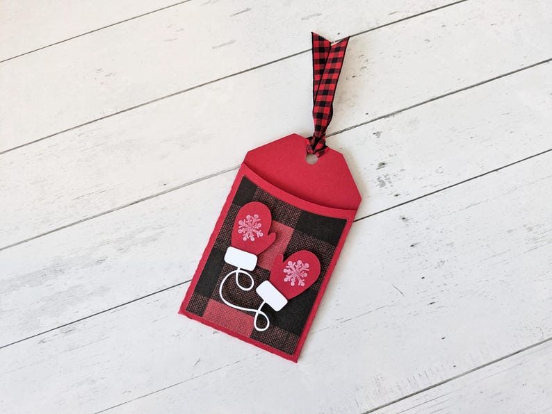 Puede incluir: Una etiqueta de regalo roja con un patr&oacute;n de cuadros negros y rojos y dos mitones rojos con detalles de copos de nieve blancos. La etiqueta tiene una cinta a cuadros roja y negra. Sobre una superficie de madera blanca.