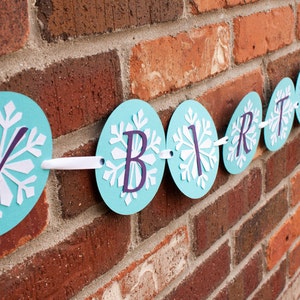 K&ouml;nnte beinhalten: Ein blaues und wei&szlig;es Geburtstagsbanner mit Schneeflockenmustern und dem Wort "Birthday" in lila Buchstaben.