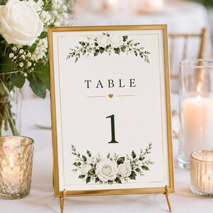 May include: A rectangular table number sign in a thin gold metal frame. The white card features the text TABLE 1 with a small gold heart icon. Floral illustrations of white roses and green leaves are printed at the top and bottom of the card.