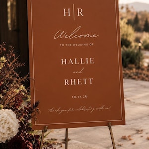 May include: A rectangular terracotta-colored wedding welcome sign featuring white text that reads H R, Welcome to the wedding of Hallie and Rhett, 10.17.26, and Thank you for celebrating with us. The sign has a thin white border and stands on a metal easel.