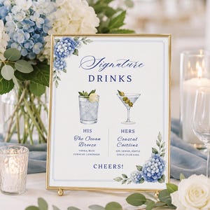 May include: A rectangular gold-framed sign featuring blue hydrangea illustrations, titled Signature DRINKS. It lists HIS The Ocean Breeze with vodka, blue curacao, lemonade and HERS Coastal Caitlins with gin, lemon, simple syrup, club soda, ending with CHEERS!