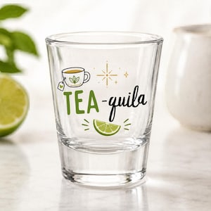 May include: Clear shot glass with the words "TEA-quila" in black script and green block letters. The glass features a tea cup graphic, a lime wedge, and a starburst design. A lime half is visible in the background.