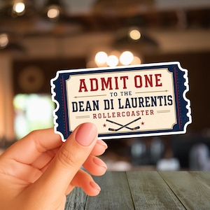 May include: A rectangular sticker shaped like an admission ticket with a cream-colored center and dark blue borders. The text reads ADMIT ONE TO THE DEAN DI LAURENTIS ROLLERCOASTER in red and blue lettering, featuring two crossed hockey sticks.