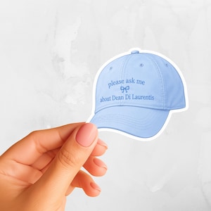 May include: A light blue baseball cap featuring the embroidered text "please ask me about Dean Di Laurentis" in a matching blue thread, centered above a small blue bow icon on the front panel of the six-panel structured crown.
