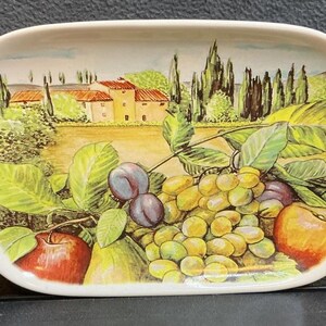 Puede incluir: Una bandeja ovalada de cerámica con una escena paisajística pintada de una villa rodeada de cipreses, con un arreglo de frutas frescas en primer plano que incluye manzanas rojas, ciruelas moradas, peras verdes y uvas verdes.
