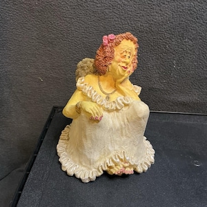May include: A figurine of a person with curly reddish-brown hair adorned with a pink flower, wearing a white ruffled dress with a textured finish. The figure has a yellow complexion, a gold-toned necklace, and rests their chin on one hand.