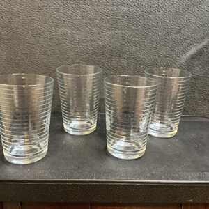 May include: A set of four clear glass tumblers, each featuring a series of thin, horizontal etched lines encircling the exterior. The glasses have a straight-sided, cylindrical shape with a slightly wider rim and a flat, circular base.