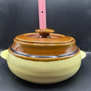 May include: A round ceramic casserole dish with a lid, featuring a two-tone design with a cream-colored base and a dark brown upper section. A pink ruler is positioned vertically behind the lid, showing a height measurement of approximately 6 inches.