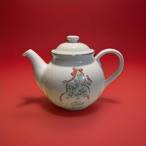May include: A cream-colored ceramic teapot with a matching lid, featuring a decorative illustration of a green ivy topiary in a pot, accented by a red ribbon bow and a small red bird perched on the side, finished with a thin green rim.