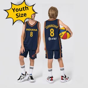 Puede incluir: Un conjunto de baloncesto azul marino con ribetes amarillos. La parte delantera muestra el texto INDIANA y el número 8. La parte trasera presenta CUNNINGHAM, el número 8 y un logotipo de AT&T. Un gráfico amarillo indica Youth Size.