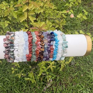 May include: A collection of multiple stretch bracelets made from various tumbled gemstone chips in a spectrum of colors including red, blue, green, purple, brown, white, and turquoise, arranged side by side on a white cylindrical display stand.