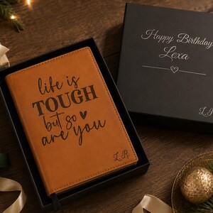 May include: A tan leather journal with the text "life is TOUGH but so are you" in a black box, next to a black box with "Happy Birthday Lexa" and a heart. The journal and box are surrounded by wrapped gifts.