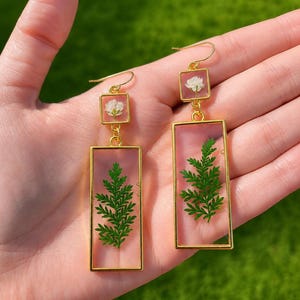 May include: A pair of gold-toned drop earrings featuring a rectangular frame and a smaller square frame. Each piece contains clear resin encasing a pressed green fern leaf and a small white flower, suspended from a gold-toned ear wire hook.