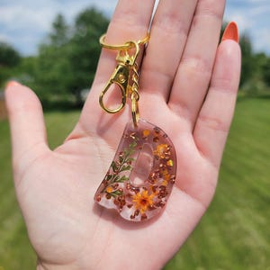 May include: A clear resin keychain in the shape of the letter D, featuring embedded dried orange flowers, green foliage, and copper-colored metallic flakes. The charm is attached to a gold-toned metal split ring and a lobster claw clasp.