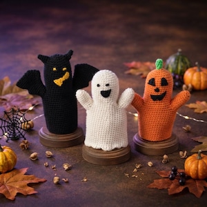 May include: Three crocheted finger puppets standing on small wooden circular bases. The left puppet is a black bat with yellow eyes and mouth. The center puppet is a white ghost with black eyes and mouth. The right puppet is an orange pumpkin.