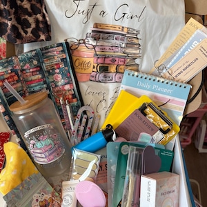 May include: A gift box containing a white drawstring bag with a stack of books and coffee illustration, a glass tumbler with a dragon graphic, a daily planner, a yellow book review notepad, skincare products, a pink heart-shaped sponge, and stationery.