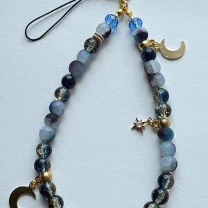 May include: A phone charm featuring a loop of blue, grey, and black beads with gold-tone accents. The charm includes two gold-tone crescent moon pendants and a small star charm with a clear stone. The text Noctis Azur is visible at the top.