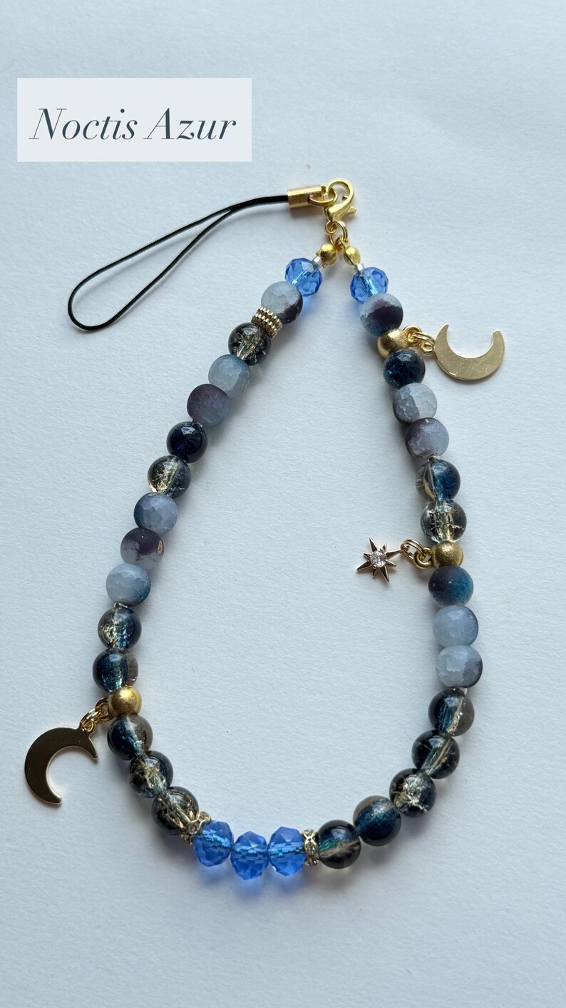 May include: A phone charm featuring a loop of blue, grey, and black beads with gold-tone accents. The charm includes two gold-tone crescent moon pendants and a small star charm with a clear stone. The text Noctis Azur is visible at the top.