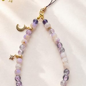 May include: A phone charm featuring a loop of purple, lavender, and clear crackle glass beads accented with gold-toned metal spacers. The accessory includes two gold-toned crescent moon charms and one small star charm with a central clear crystal.
