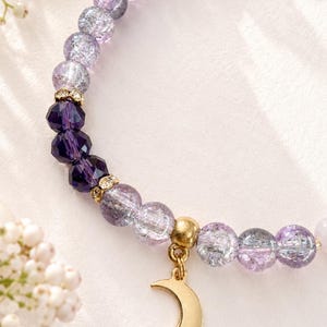May include: A beaded bracelet featuring light purple crackle glass beads, deep purple faceted crystal beads, and gold-toned spacers. The bracelet includes a gold-toned crescent moon charm and a small gold-toned star charm with a central crystal.