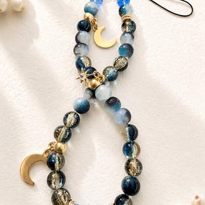 May include: A beaded phone charm featuring a loop of blue, navy, and clear glass beads with gold-toned accents. The charm includes a gold-colored crescent moon pendant and a small star-shaped charm, finished with a black cord attachment.