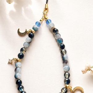 May include: A phone charm featuring a loop of blue, navy, and translucent beads in varying shades and textures. The accessory includes gold-toned metal charms shaped like two crescent moons and a star, with a black cord attachment at the top.