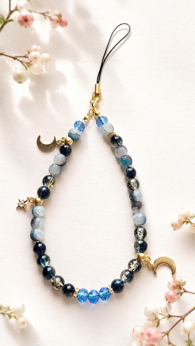 May include: A phone charm featuring a loop of blue, navy, and translucent beads in varying shades and textures. The accessory includes gold-toned metal charms shaped like two crescent moons and a star, with a black cord attachment at the top.