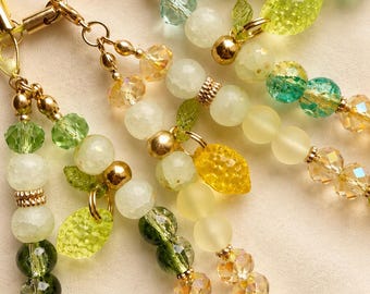May include: Close-up of several beaded phone charms in shades of green, yellow, and clear. The charms feature a variety of beads, including faceted crystals, round beads, and leaf-shaped charms. Gold-colored metal accents and clasps complete the design.