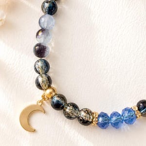 May include: A beaded bracelet featuring a mix of round, dark blue, and translucent grey beads with gold-toned accents. A gold-toned crescent moon charm hangs from the bracelet, which also includes three faceted, bright blue crystal beads.