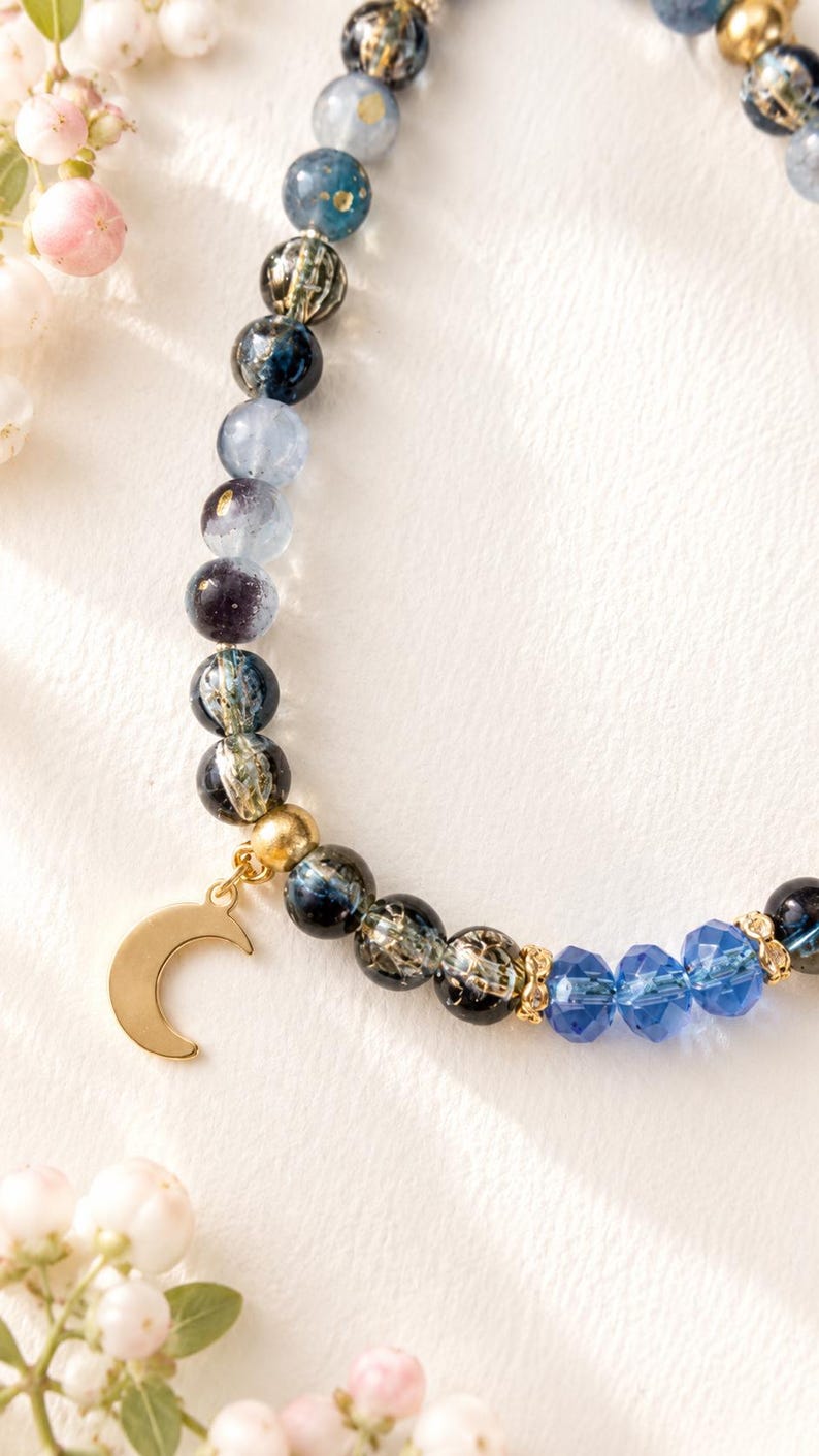 May include: A beaded bracelet featuring a mix of round, dark blue, and translucent grey beads with gold-toned accents. A gold-toned crescent moon charm hangs from the bracelet, which also includes three faceted, bright blue crystal beads.