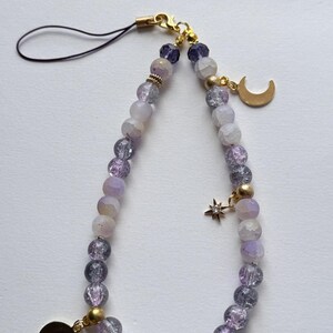 May include: A phone charm featuring a loop of purple and white crackle glass beads, accented with gold-toned metal spacers. The accessory includes two gold-colored crescent moon charms and a small star charm with a clear stone, finished with a black cord.