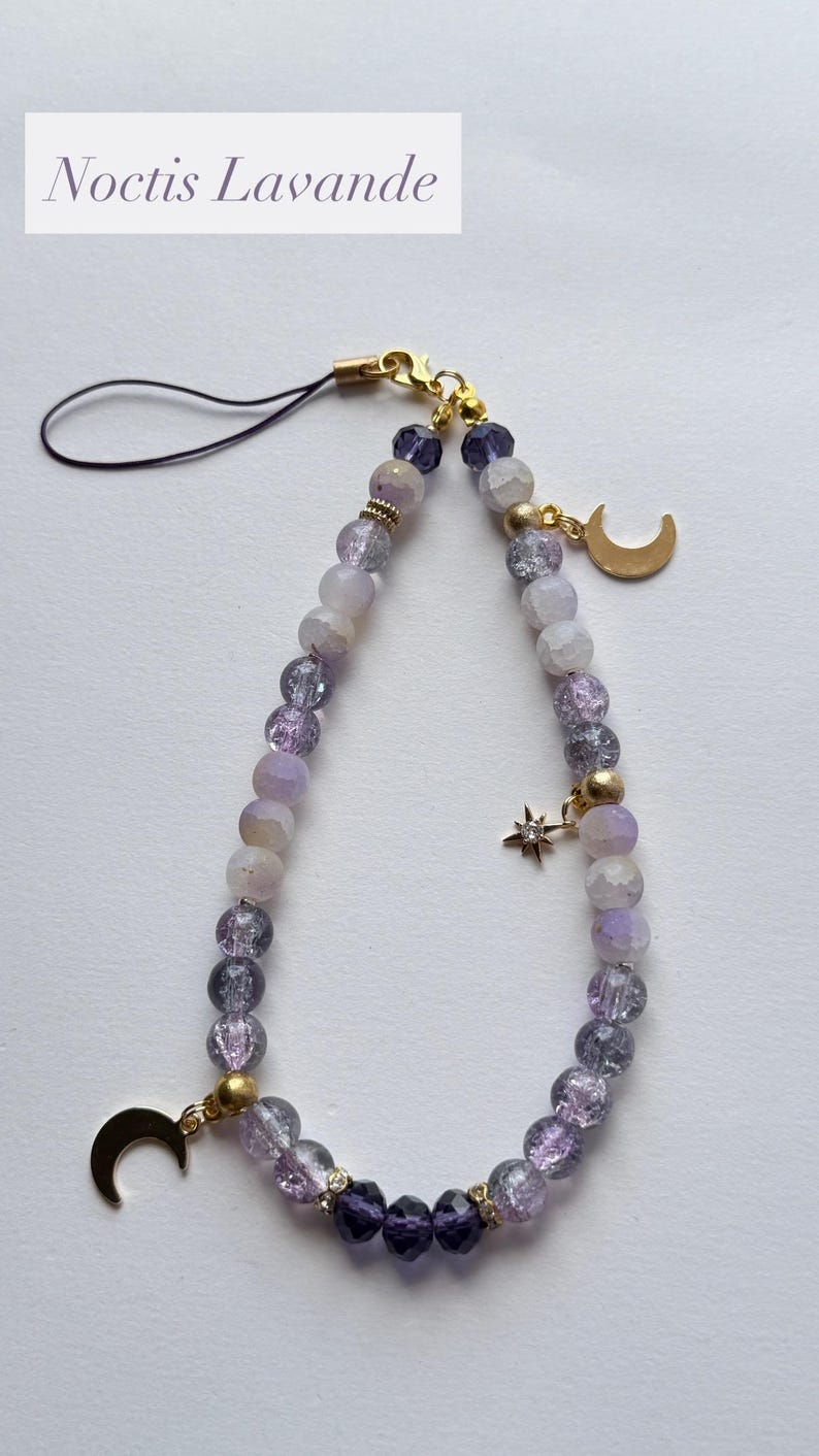 May include: A phone charm featuring a loop of purple and white crackle glass beads, accented with gold-toned metal spacers. The accessory includes two gold-colored crescent moon charms and a small star charm with a clear stone, finished with a black cord.