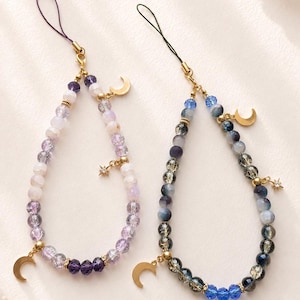 May include: Two beaded phone charms featuring faceted glass beads in shades of purple and blue. Each charm includes a gold-toned crescent moon pendant, a small star charm, and a thin cord loop for attachment to a mobile device or accessory.
