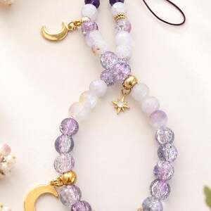 May include: A phone charm featuring a loop of purple and white beads in varying shades and textures. The charm includes gold-toned metal accents, a crescent moon pendant, a small star charm with a clear stone, and a dark purple cord attachment.