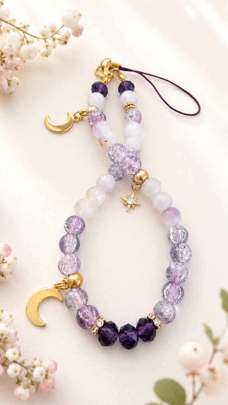 May include: A phone charm featuring a loop of purple and white beads in varying shades and textures. The charm includes gold-toned metal accents, a crescent moon pendant, a small star charm with a clear stone, and a dark purple cord attachment.