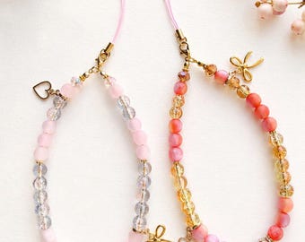 May include: Two beaded phone charms with pink string loops. The left charm features matte pink and clear faceted beads with a gold heart charm. The right charm features coral, pink, and amber faceted beads with a gold bow charm and gold heart.